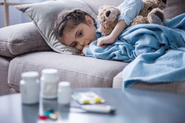 毛布で覆われて病気の小さな女の子がテディベアを抱いてとソファの上に横たわっている間医学の悲しいことに探して - 写真・画像