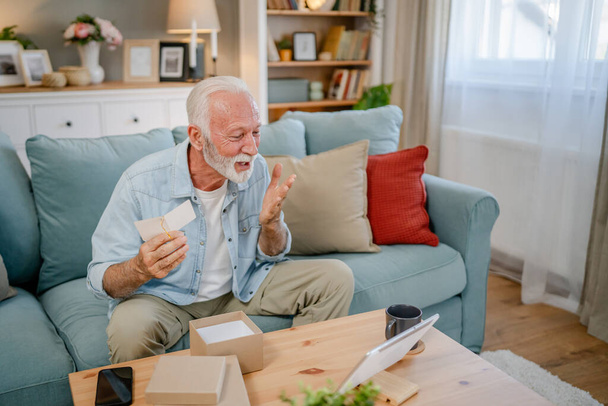 Ένας ηλικιωμένος συνταξιούχος παππούς με γκρίζα μαλλιά και γενειάδα λαμβάνουν δώρα σε κουτί ανοιχτή ανάγνωση κάρτα χαρούμενο χαμόγελο μπροστά από φορητό υπολογιστή στο σπίτι έχοντας online video call copy space - Φωτογραφία, εικόνα