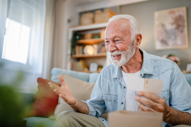 Bir son sınıf büyükbaba emeklisi gri saçlı ve sakallı hediye alıyor açık kutuda kart okuyarak mutlu bir gülümseme evde dizüstü bilgisayarın önünde internetten fotokopi odası arıyor. - Fotoğraf, Görsel
