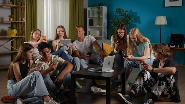 Foto full-size di un gruppo di adolescenti, giovani, amici seduti su un divano e sul pavimento intorno al tavolo, usando i loro smartphone, laptop, tablet, chattare, parlare. Pubblicità, gen z, moderno - Foto, immagini