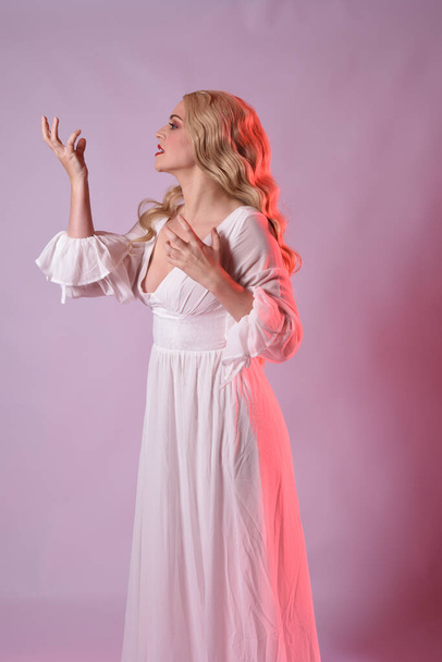 Close up portrait of beautiful blonde model wearing elegant  white halloween gown. arms reaching out like hungry vampire.  isolated on studio background with  red cinematic moody lighting. - Foto, Imagem