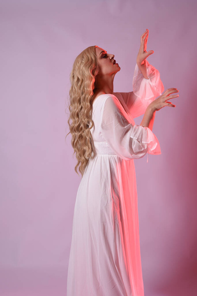 Detailní portrét krásné blond modelky v elegantních bílých halloween šatech. paže natahují jako hladový upír. izolované na pozadí studia s červeným kinematografickým náladovým osvětlením. - Fotografie, Obrázek