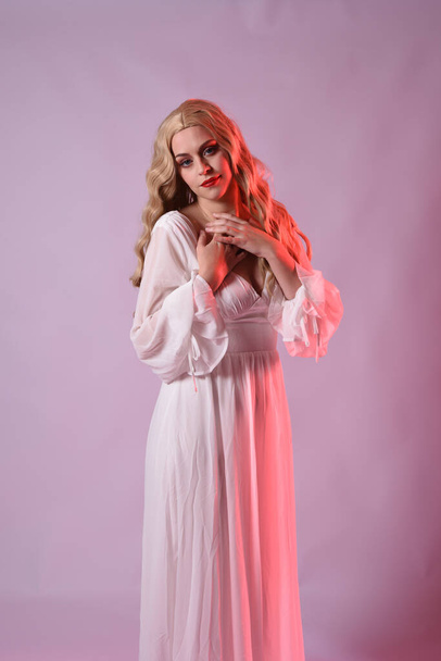 Close up portrait of beautiful blonde model wearing elegant  white halloween gown. arms reaching out like hungry vampire.  isolated on studio background with  red cinematic moody lighting. - Photo, Image