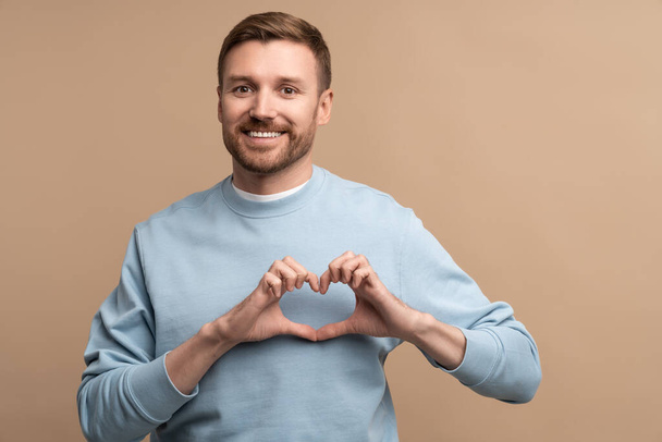 Romantic guy making heart shape figure with fingers, isolated on studio beige background. Joyful man feeling happy show love symbol looking at camera. Inspired male with love sign for advertising. - Photo, Image