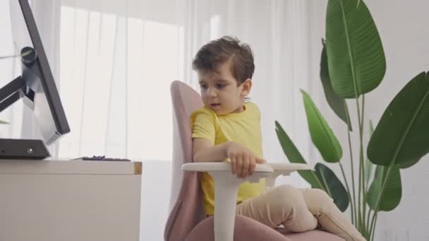 小さな子供の男の子は、オンラインレッスンを受けた後、ラップトップコンピュータの前で快適なファッショナブルな回転椅子にリラックスします - 映像、動画