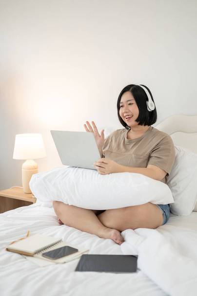 Szczęśliwa i czarująca Azjatka plus-size rozmawiająca z przyjaciółmi przez laptopa, siedząc na łóżku w sypialni. - Zdjęcie, obraz
