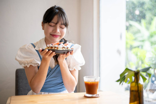 Una bella e felice giovane donna asiatica che profuma di waffle o croffle appena sfornati, godendosi il pomeriggio in una caffetteria. - Foto, immagini