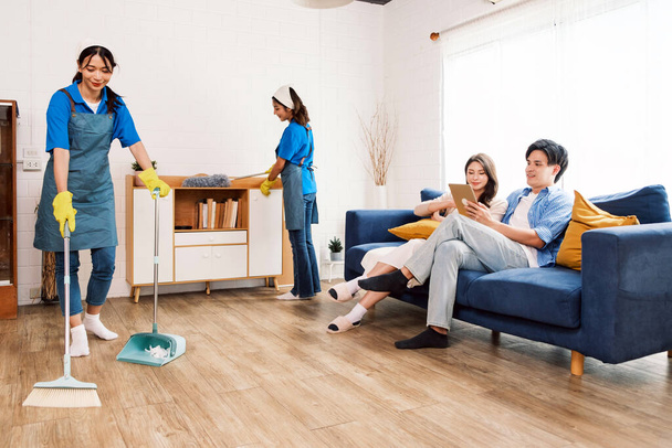Νεαρές επαγγελματίες καθαρίστριες εργαζόμενες που εργάζονται στο σπίτι. Το ένα σκουπίζει σκουπόξυλα στο ξύλινο πάτωμα και το άλλο καθαρίζει το χαμηλό ντουλάπι ενώ δύο ιδιοκτήτες κάθονται στον καναπέ.. - Φωτογραφία, εικόνα