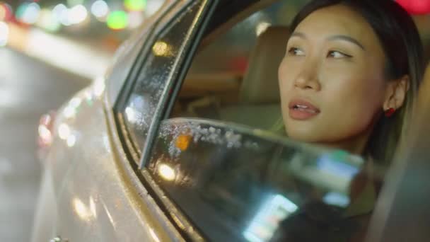 Uitzicht van buiten de auto van jonge Aziatische vrouw op zoek naar 's nachts stad door middel van geopende venster en het nemen van telefoongesprek tijdens het rijden op de achterbank - Video