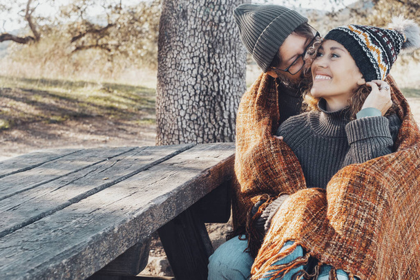 Glückliches Paar in romantischer Freizeitaktivität zusammen, beim Kuscheln im Park. Herbst liebt Menschen. Mann umarmt und klebt, niedliche Frau sitzt auf einer Holzbank mit gelben Blättern und Bäumen im Hintergrund - Foto, Bild
