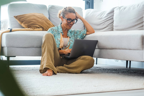 Tasasız, modern, yetişkin bir kadın evde dizüstü bilgisayar kullanıyor gri bir kanepeye karşı yerde oturuyor. İnternette tek başına bilgisayarda sörf yapan bir kadın eğleniyor. Çevrimiçi aktivite boş zaman kapalı alanda - Fotoğraf, Görsel