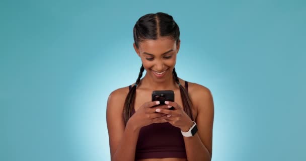 Telefoon, glimlach en vrouw in een studio netwerken op sociale media, mobiele app of het internet. Happy, technologie en jonge Indiase vrouwelijke model scroll op website met mobiele telefoon geïsoleerd door blauwe achtergrond. - Video