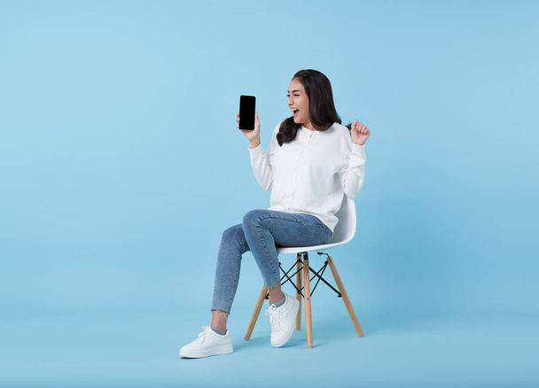 Felice giovane donna asiatica mostrando smartphone a schermo vuoto. Mentre lei seduta sulla sedia bianca isolata su sfondo blu studio. - Foto, immagini
