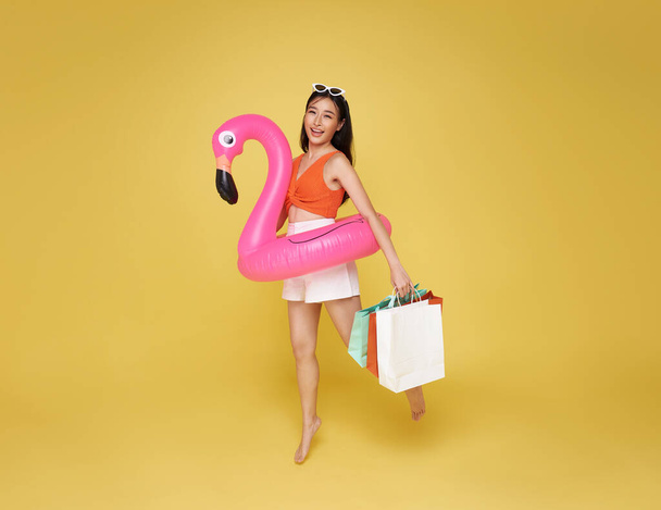 Heureuse jeune femme asiatique sexy vêtue de maillots de bain tenant sac à provisions et anneau en caoutchouc flamant rose sautant pour la vente d'été sur fond jaune. - Photo, image