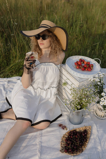 Schöne Blondine im weißen Sommerkleid, eleganter Hut sitzt auf einer weißen Decke mit Früchten und genießt die Aussicht. Konzept des Picknicks in der Natur während der Sommerferien oder am Wochenende. Das Leben genießen. - Foto, Bild