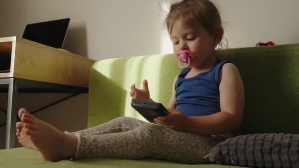 Bébé fille regardant attentivement l'écran d'une console de jeu portable, assis sur un canapé dans une pièce. Jeux vidéo, jeux mobiles, réalité virtuelle, e-sport - Séquence, vidéo