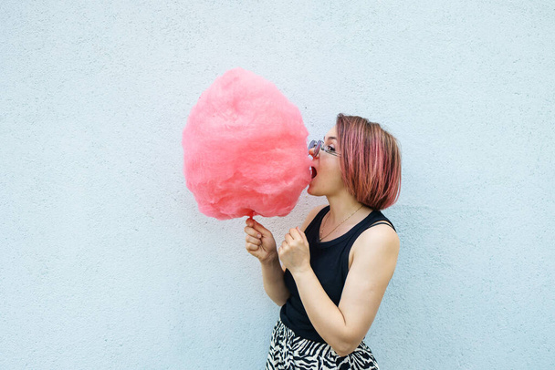 young beautiful hipster woman with colorful hair and fashion sunglasses eating cotton candy against white wall. - Photo, Image
