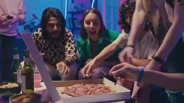 Střední záběr šťastných různorodých mladých mužů a žen sedících kolem stolu s pivem na párty, otevírající krabici od pizzy, jásající, smějící se a všichni si dávají plátek - Záběry, video