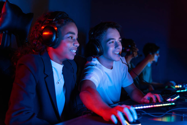 White boy and black girl teens in headsets playing video games in video game club with blue and red illumination. Keyboard and mouse with illumination - Zdjęcie, obraz