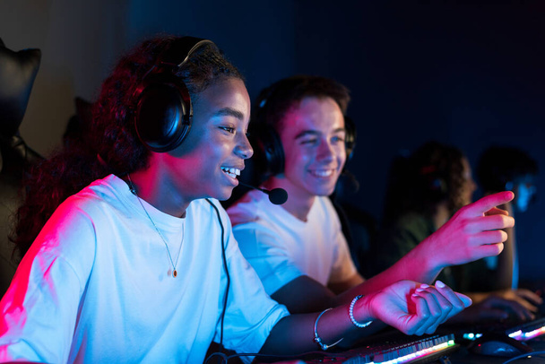 Weiße Jungen und schwarze Mädchen Teenager mit Headsets spielen Videospiele im Videospielclub mit blauer und roter Beleuchtung. Tastatur mit Beleuchtung - Foto, Bild