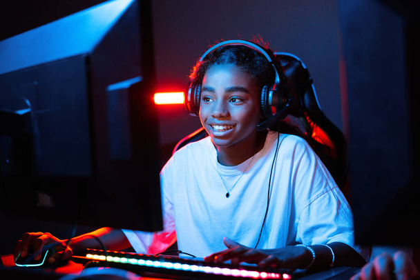Black teen smiling girl in headset playing video games in video game club with blue and red illumination. Keyboard with illumination - Фото, зображення