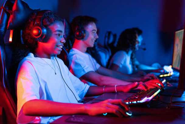 Group of multiracial teens in headsets playing video games in video game club with blue and red illumination. Keyboard and mouse with illumination - Foto, Imagen