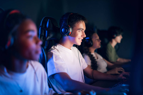 Tiener lachende jongen in headset spelen van video games in video game club met blauwe en rode verlichting - Foto, afbeelding