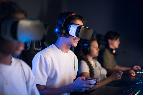 Tiener lachende jongen in VR headset spelen van video games in video game club met blauwe verlichting met behulp van een gamepad - Foto, afbeelding