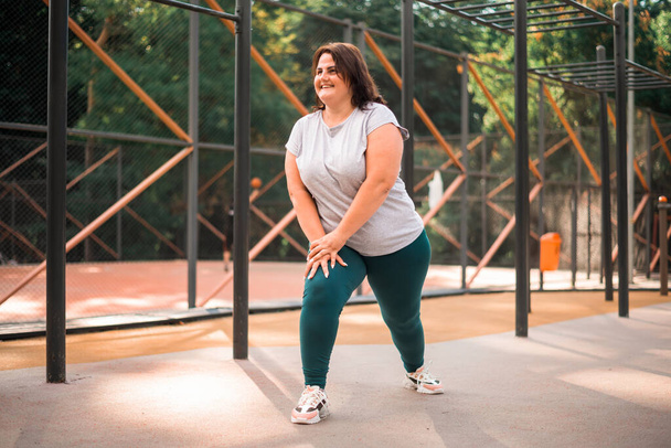 Χαμογελαστή γυναίκα με υπέρβαρη φόρμα να κάνει ασκήσεις σε γήπεδο σε πάρκο. - Φωτογραφία, εικόνα