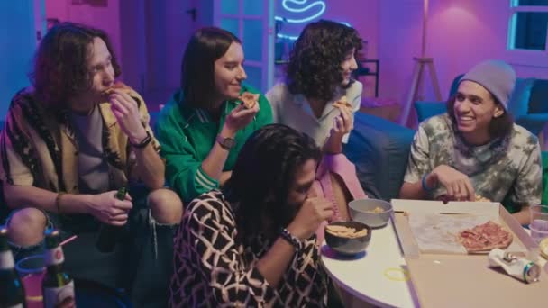 Střední záběr pěti multietnických holek a chlapů sedících na gauči u stolu v pokoji, jedoucích pizzu, bramborové lupínky, pijících pivo, sledujících komediální show v televizi, klábosících a ukazujících prstem - Záběry, video