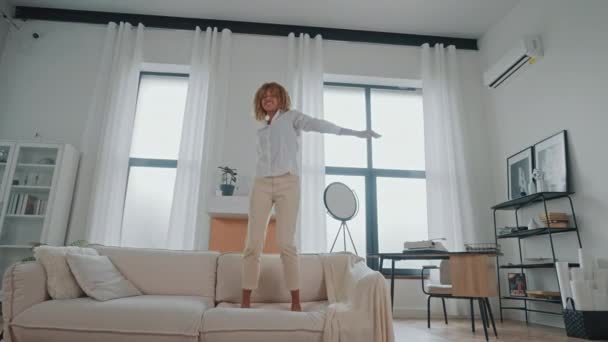 mujer excitada con el pelo rubio usando ropa casual saltando en el sofá en casa, mostrando emociones positivas - Metraje, vídeo