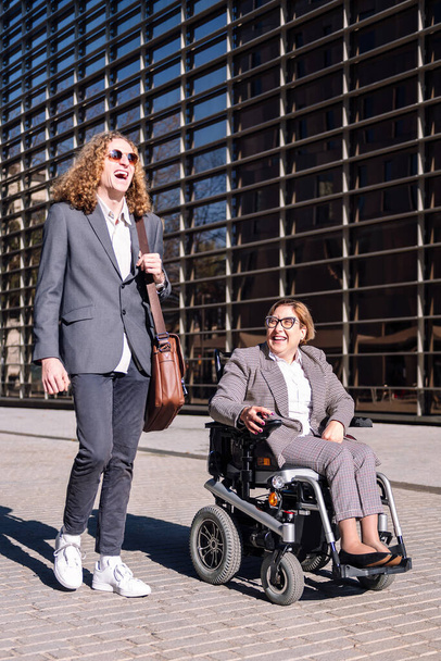 Geschäftsmann und Frau im Rollstuhl lachen glücklich zwischen Bürogebäuden im Finanzviertel, Konzept der Vielfalt und des urbanen Lebensstils, Kopierraum für Text - Foto, Bild
