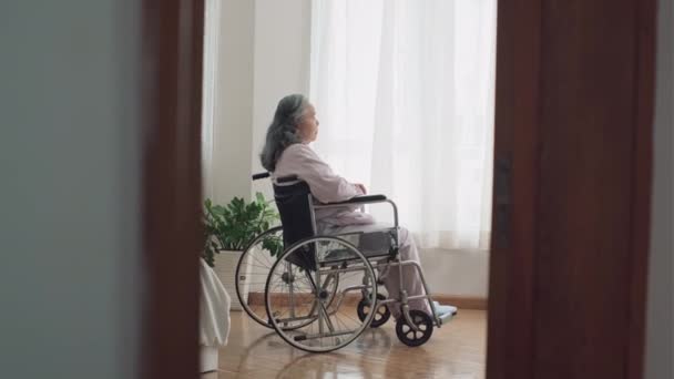 Długie ujęcie przygnębionej starszej kobiety niepełnosprawnej na wózku inwalidzkim patrzącej przez okno w domu opieki - Materiał filmowy, wideo