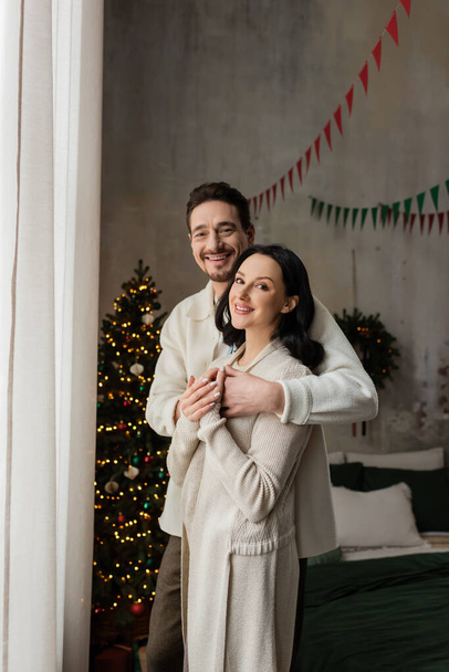 positive man hugging wife in cozy home wear near blurred decorated Christmas tree, winter holidays - 写真・画像