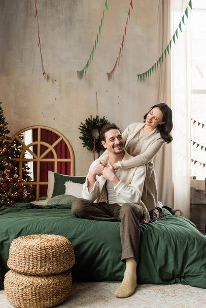 陽気な結婚したカップルは,現代のアパートで装飾されたクリスマスツリーの近くのベッドに座って抱擁し, - 写真・画像