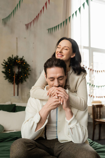 starostlivý muž sedí na posteli a líbání rukou šťastné ženy v blízkosti vánoční věnec v moderním bytě - Fotografie, Obrázek