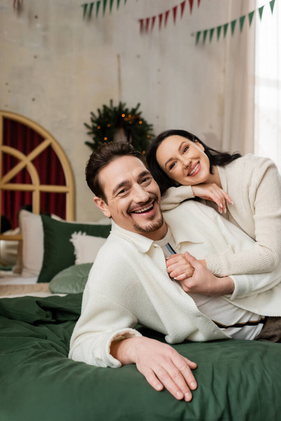 σύζυγος αγκαλιάζει χαρούμενη σύζυγο και αναπαύεται μαζί στο κρεβάτι κοντά στο θολό στεφάνι των Χριστουγέννων στον τοίχο - Φωτογραφία, εικόνα