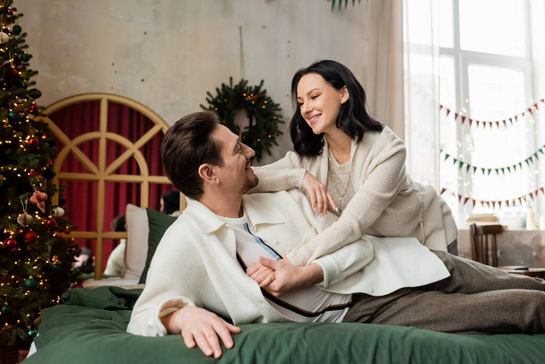 husband embracing cheerful wife and lying on bed near decorated Christmas tree and wreath on wall - 写真・画像