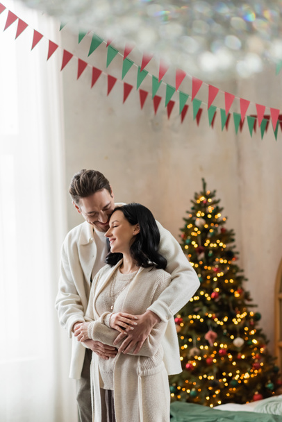 fröhliches Ehepaar im Haus umarmt sich und steht gemeinsam neben verschwommenem Weihnachtsbaum - Foto, Bild