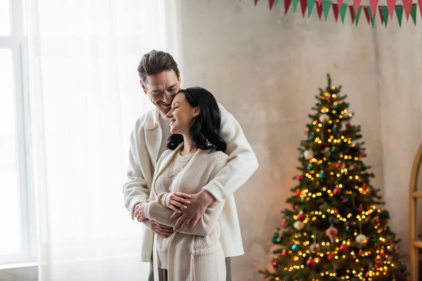 joyful and married couple in home wear hugging and standing together near blurred Christmas tree - Photo, Image