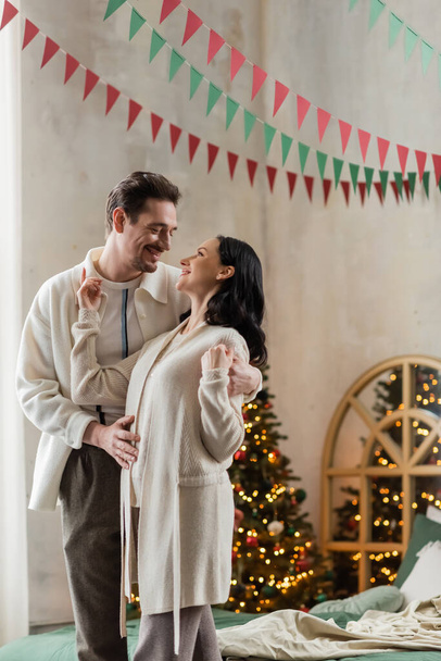 joyful husband and wife in home wear hugging and standing together near blurred Christmas tree - Photo, Image