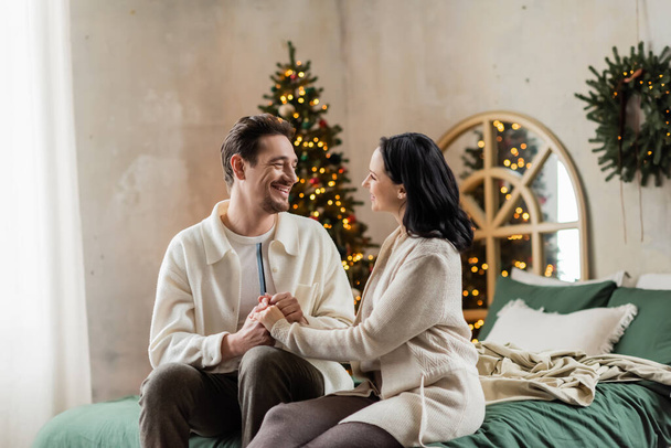 joyful husband and wife in home wear sitting on bed and holding hands near blurred Christmas tree - Φωτογραφία, εικόνα