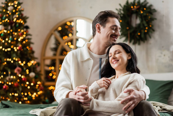 happily married couple embracing each other near blurred lights of Christmas tree on backdrop, cozy - Photo, Image