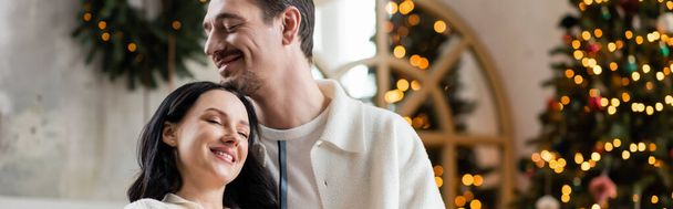 happily married couple embracing near blurred lights of Christmas tree on backdrop, cozy banner - 写真・画像