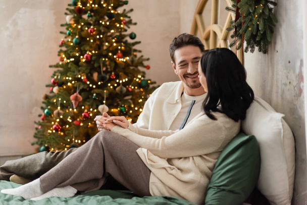 cheerful married couple spending cozy morning near blurred lights of Christmas tree on backdrop - Foto, Bild