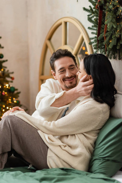 hombre alegre tocando suavemente la cara de la esposa cerca de las luces borrosas del árbol de Navidad, mañana acogedora - Foto, Imagen
