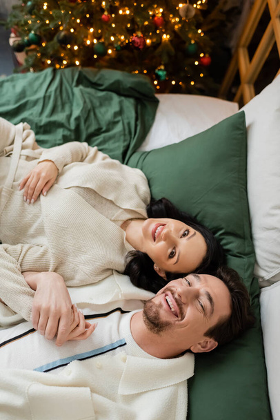 vrchol pohled na radostný pár utrácet útulné ráno a ležet v posteli v blízkosti zdobené vánoční stromeček - Fotografie, Obrázek