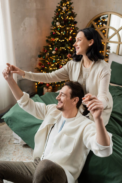 brunette woman holding hands of husband and spending time together in bedroom near Christmas tree - Photo, Image