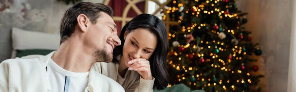 season of joy, happy man chatting with wife near decorated Christmas tree in modern bedroom, banner - Photo, Image