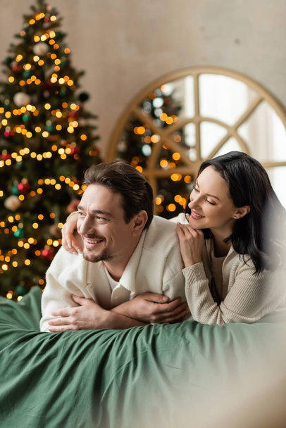 portrait of dreamy couple looking away and lying together on bed near Christmas tree with lights - Photo, Image